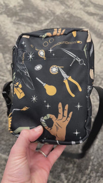 Video of a crossbody bag that is black with jewelry tools printed all over it in various colors, being held in a hand.