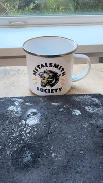 Metalsmith Society Panther Quench Cup enamel mug filled with water on a jewelry maker's soldering bench, used as a quench cup for metalsmithing.
